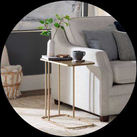 Gold side table next to a beige couch with pillows. A vase with greenery, a cup, and books are on the table.
