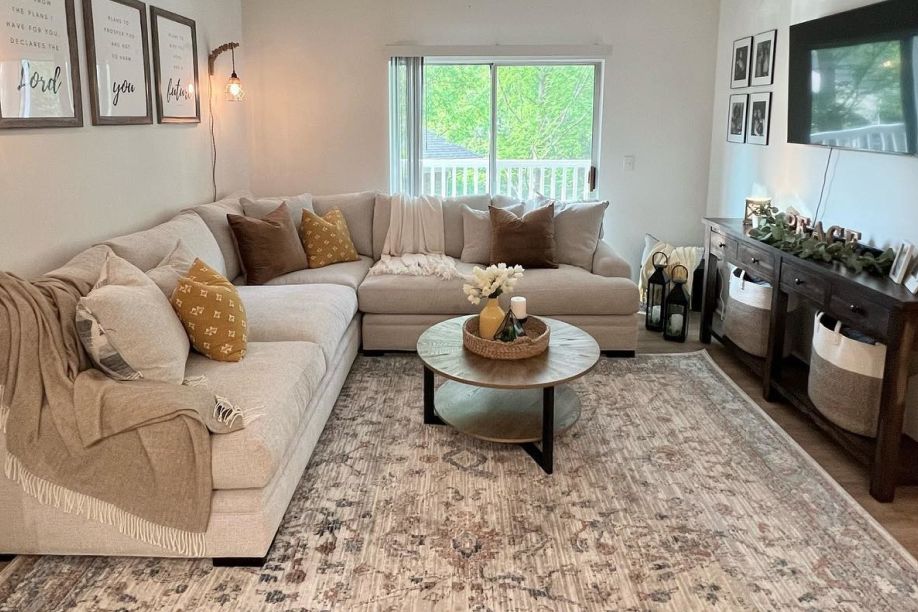 A living room with a beige sectional sofa, a round coffee table, and a patterned rug.
