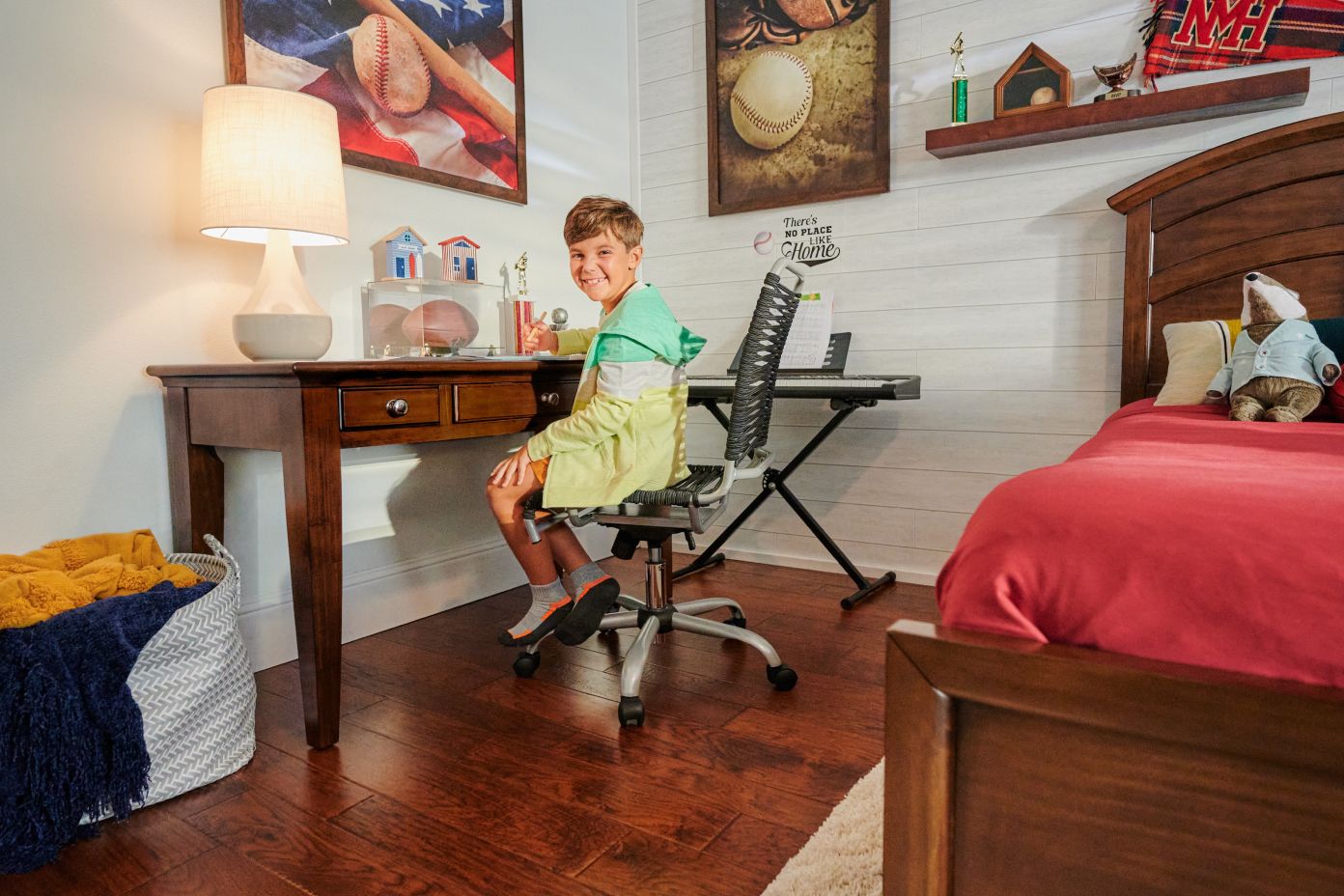 A boy sitting at a desk in a bedroom with a bed and a keyboard.
