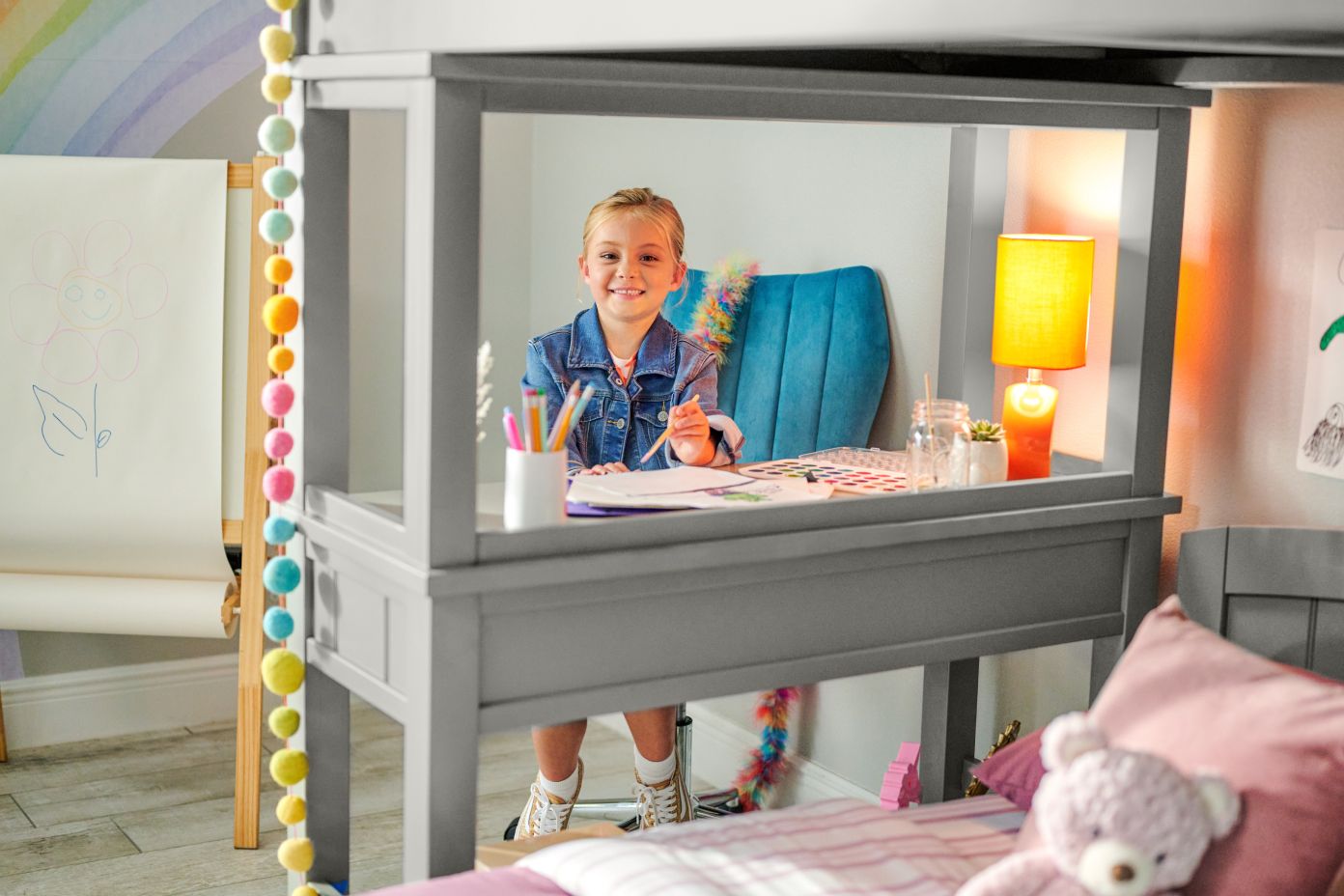 A young girl sits at a desk in a loft bed, smiling and drawing.
