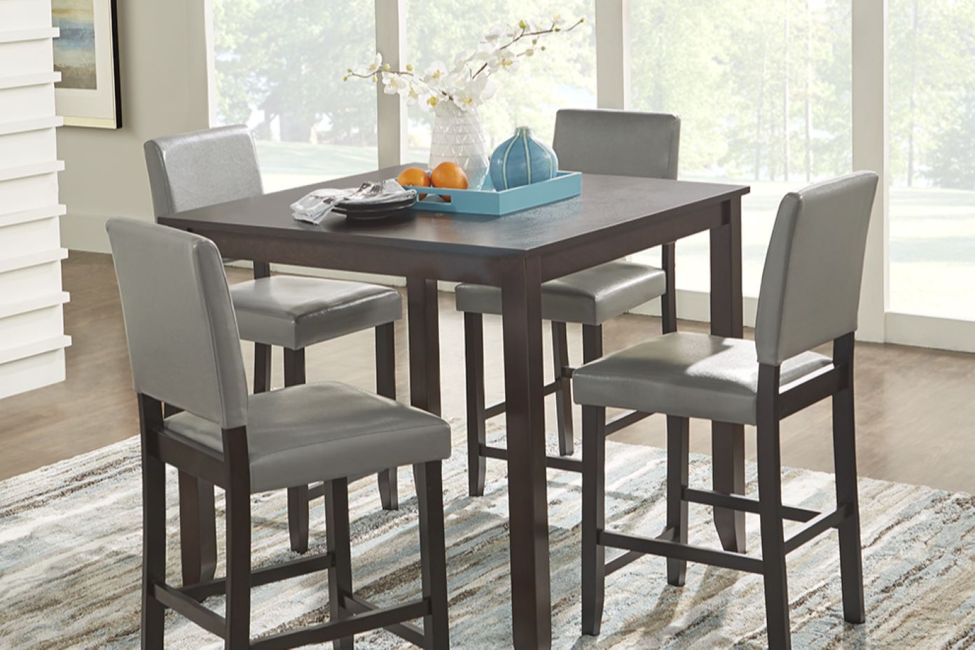 A dark brown square dining table with four gray upholstered bar stools.
