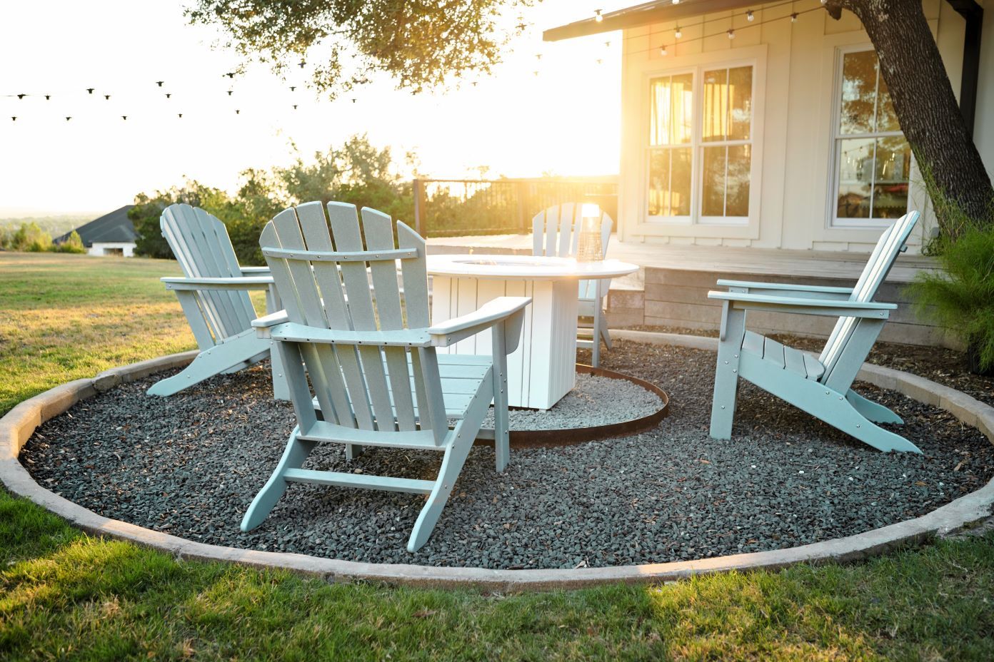 Four gray Adirondack chairs around a white fire pit on a gravel patio.
