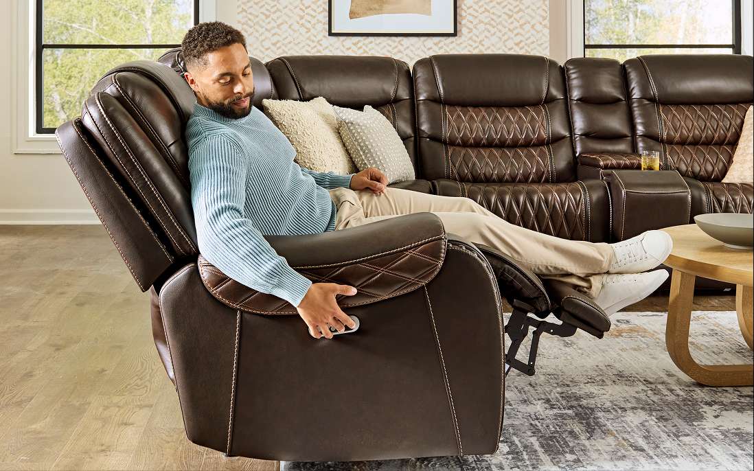 A man reclines on a brown leather recliner sofa, adjusting the controls.
