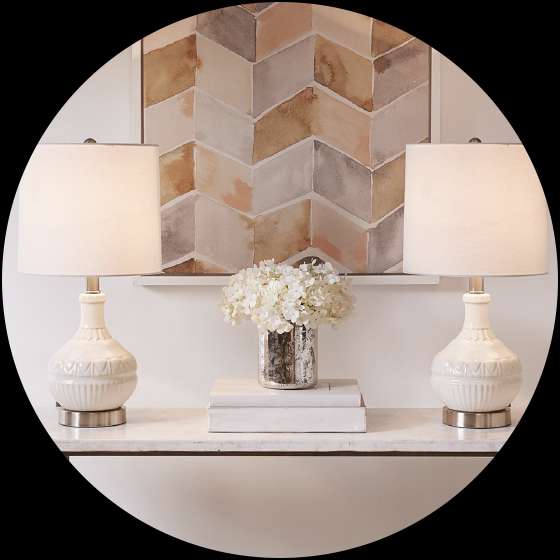 Two white lamps on a marble table with a vase of flowers and a painting in the background.
