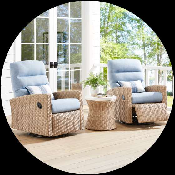 Two wicker recliner chairs with blue cushions on a wooden deck. A small side table with a plant sits between them.
