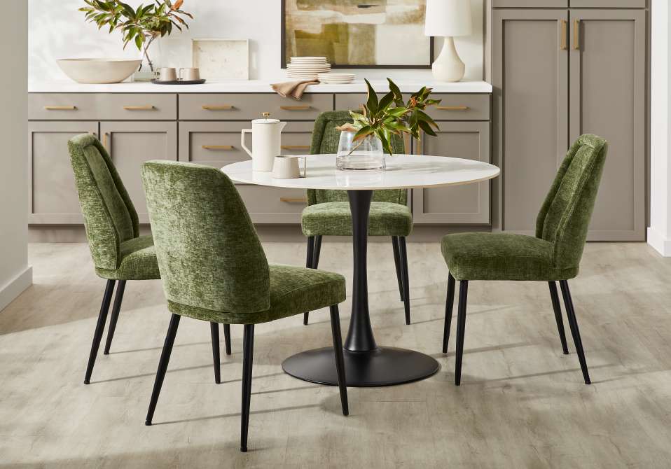 A round white dining table with four green velvet chairs.
