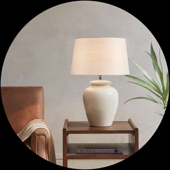 Cream-colored lamp on a wooden nightstand next to a leather chair and a plant.
