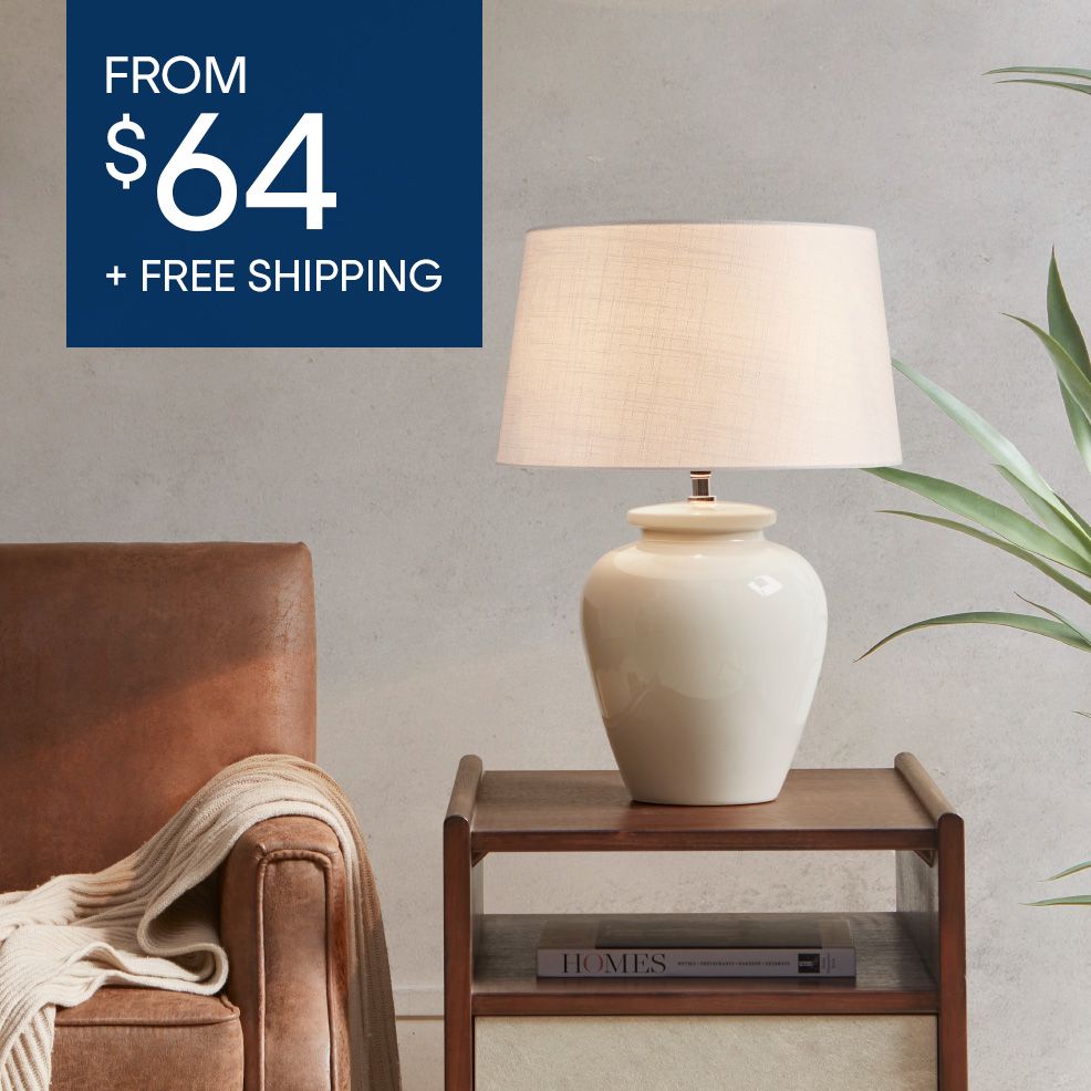 FROM $64 + FREE SHIPPING. A cream-colored lamp on a wooden nightstand.
