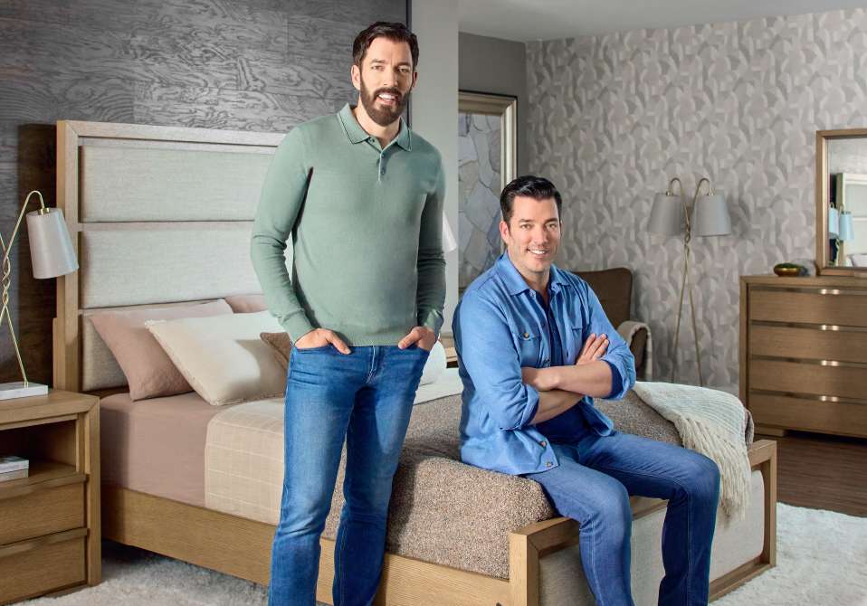 Two men in a bedroom, one standing and one sitting on the bed.
