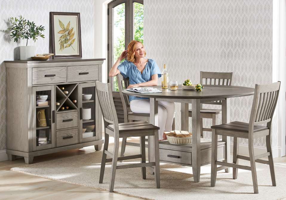 A woman sits at a round dining table with chairs. There is a buffet in the background.
