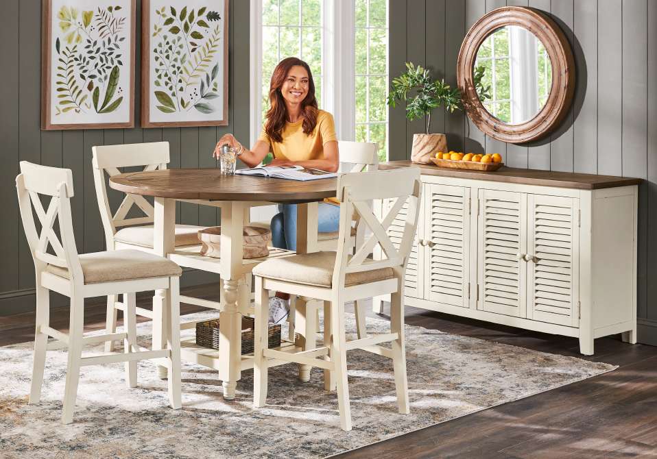 A woman sits at a round table with chairs. There is a buffet and mirror in the background.
