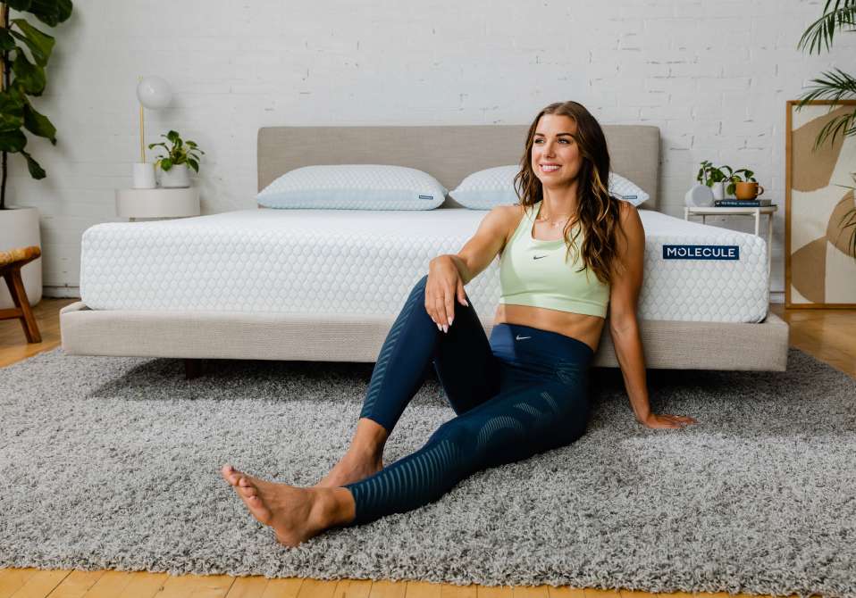 A woman in athletic wear sits on a rug in front of a bed with a Molecule mattress.
