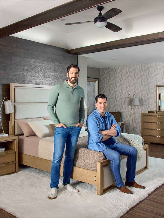 Two men in a bedroom, one standing and one sitting on a bed.
