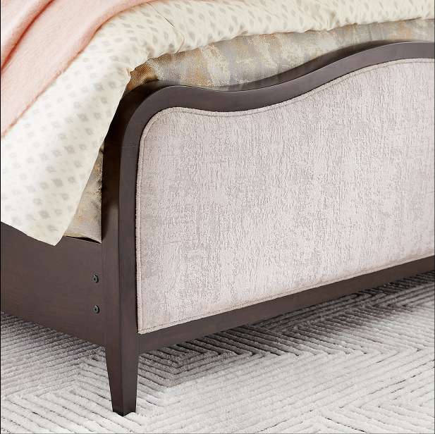 Dark brown bed frame with a light-colored upholstered headboard and a patterned rug.
