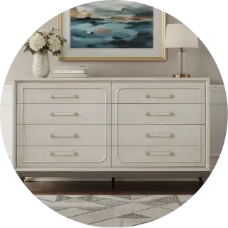 White dresser with gold hardware, topped with a painting, lamp, and flowers.
