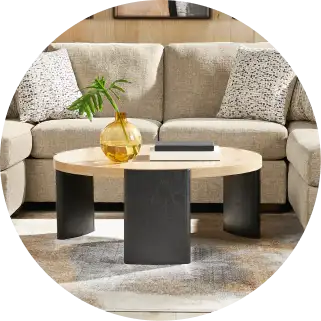 Round coffee table with a light wood top and black legs, in front of a beige sofa.
