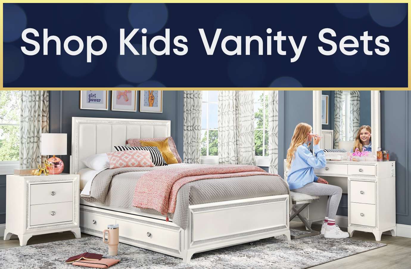 Shop Kids Vanity Sets. A bedroom with a white bed, nightstand, and vanity set. A girl is sitting at the vanity.