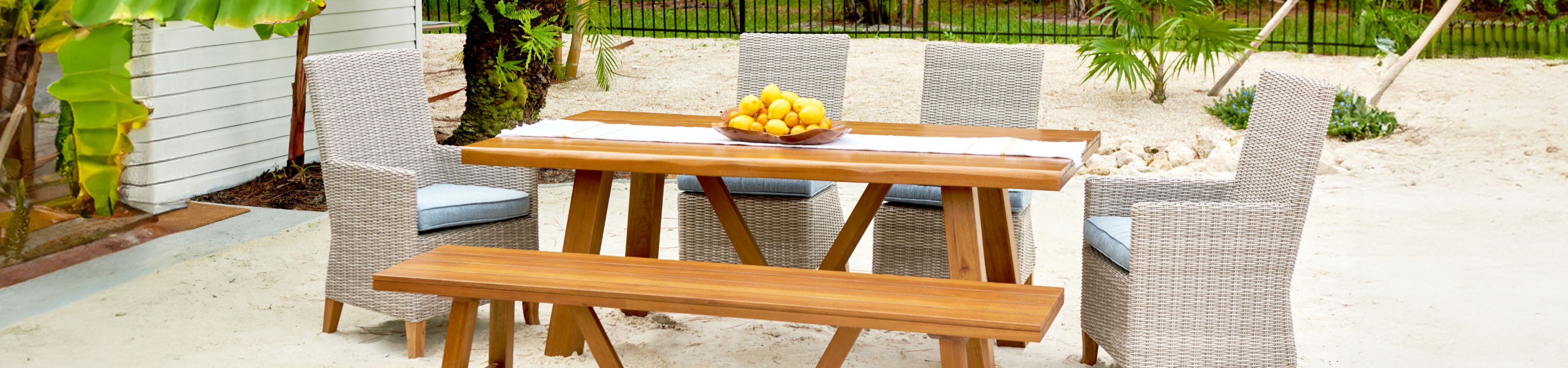 Outdoor dining set with a wooden table, bench, and woven chairs. A bowl of lemons sits on the table.
