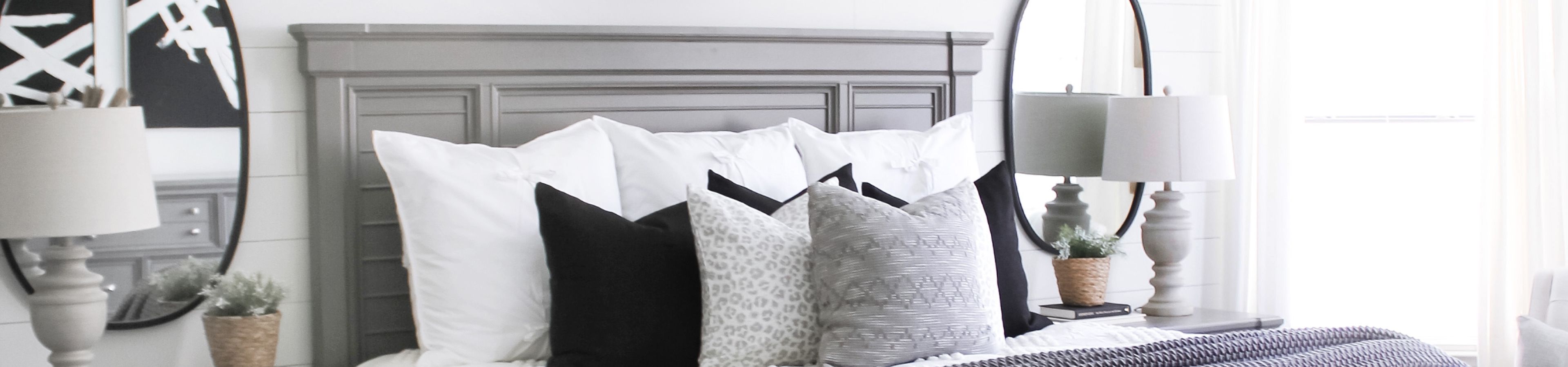 Bedroom with a gray headboard, white bedding, and decorative pillows. Two lamps and a mirror are on the side table.
