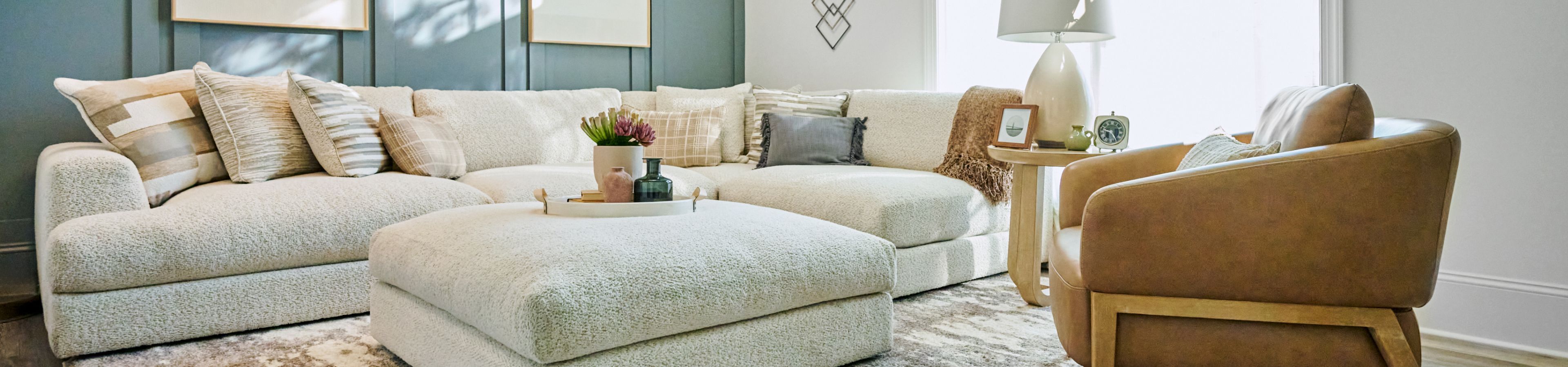 A living room with a cream-colored sectional sofa, ottoman, and a brown leather chair.
