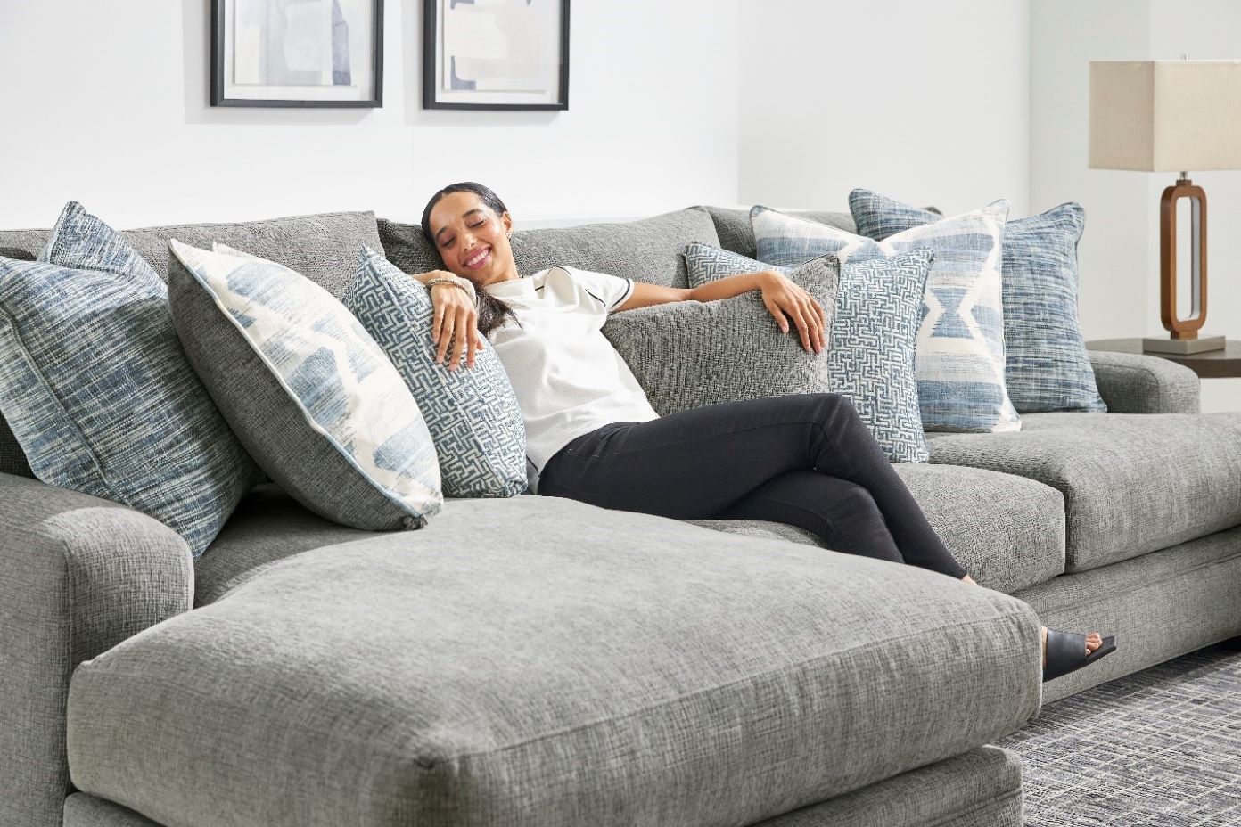 A woman relaxes on a gray sectional sofa with blue patterned pillows.
