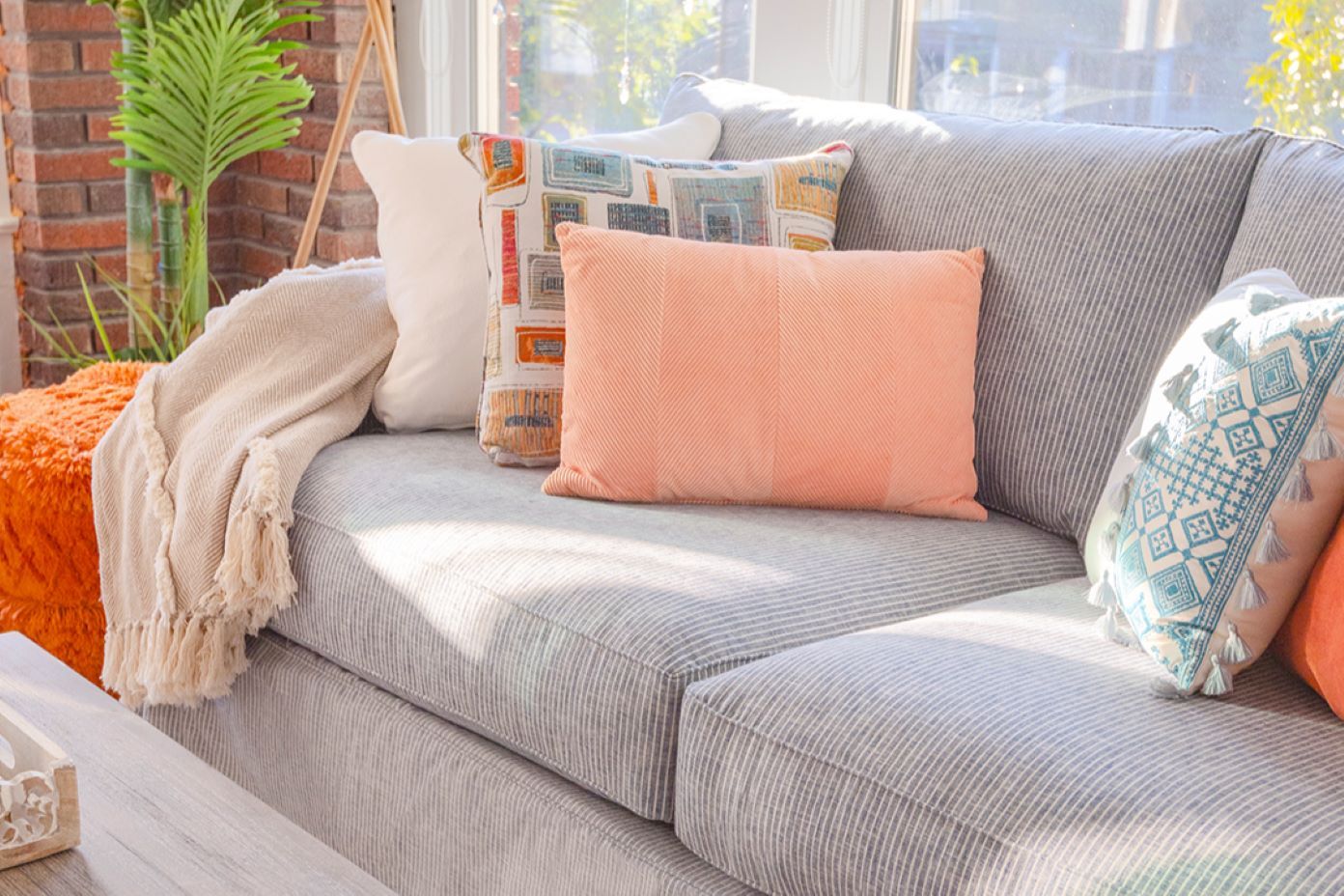 A gray striped couch with several decorative pillows. A blanket and orange ottoman are also visible.
