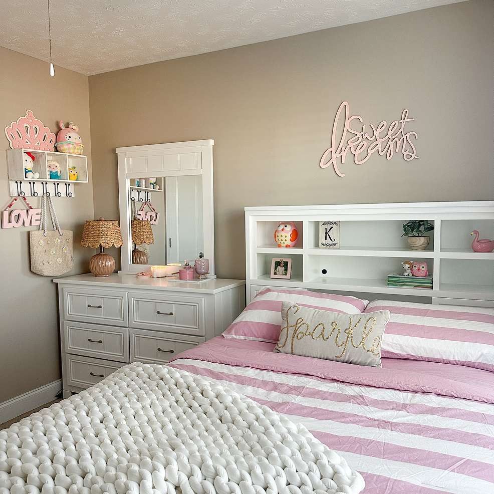 A bedroom with a white dresser, bed, and pink and white striped bedding. A "Sweet Dreams" sign is on the wall.
