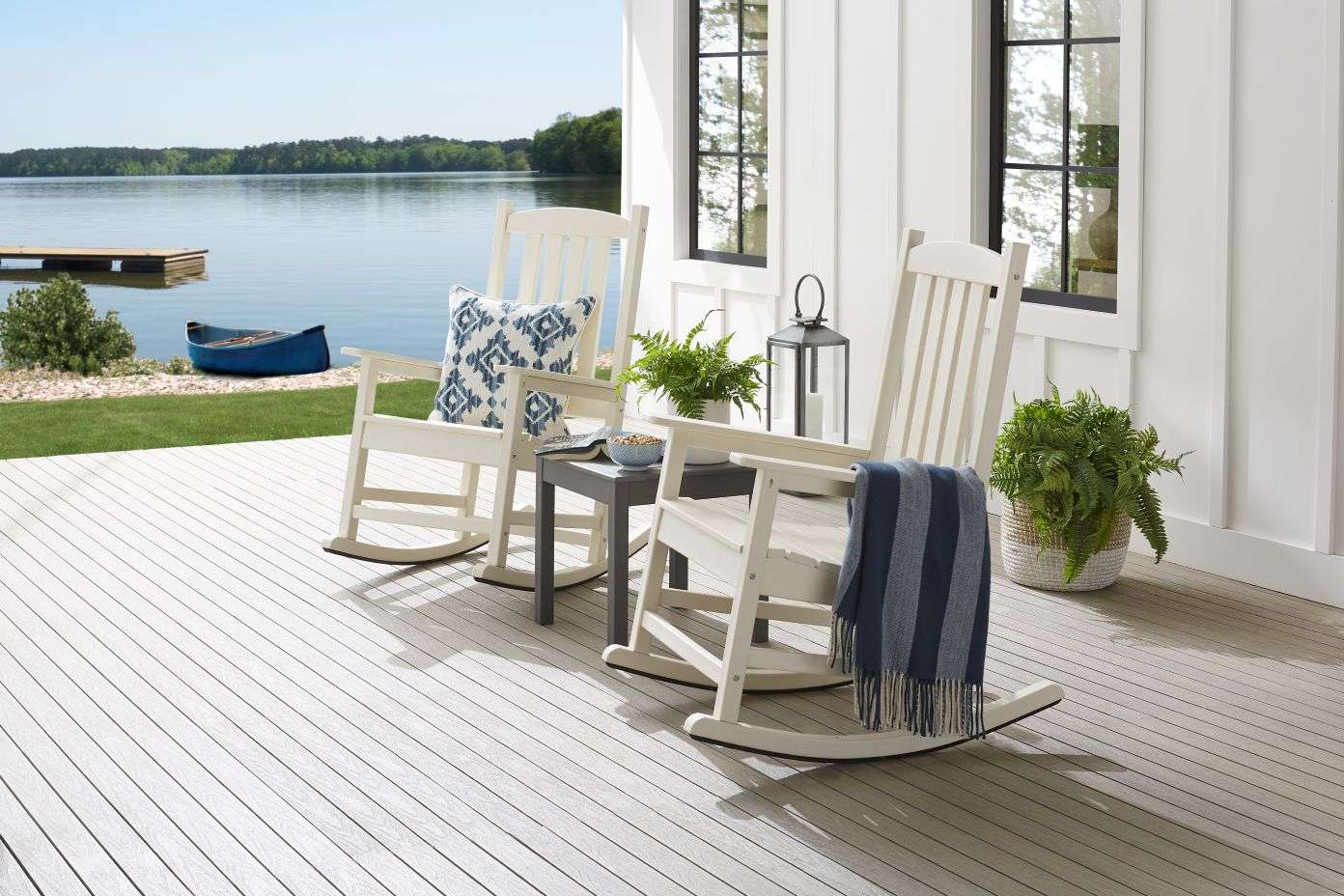 Two white rocking chairs on a deck overlooking a lake. A blue blanket and pillow are on the chairs.