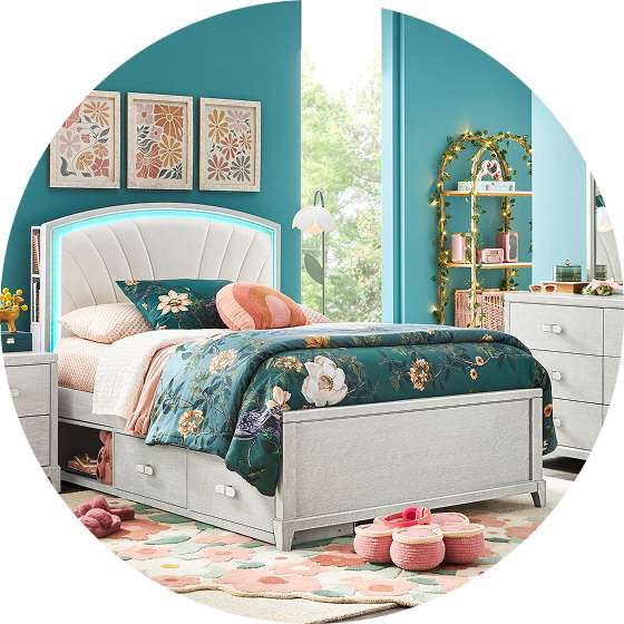 A bedroom with a bed, nightstand, and dresser. The bed has a floral comforter and a headboard with a light.
