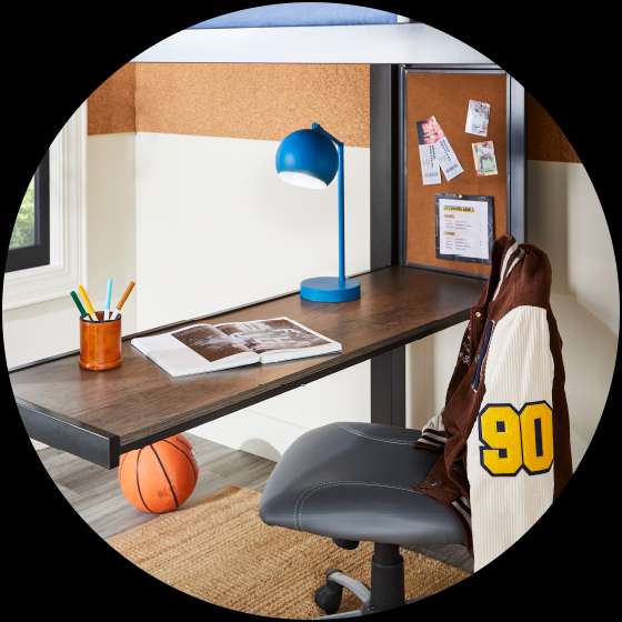 A loft bed with a desk, a blue lamp, and a basketball. A jacket hangs on the chair.
