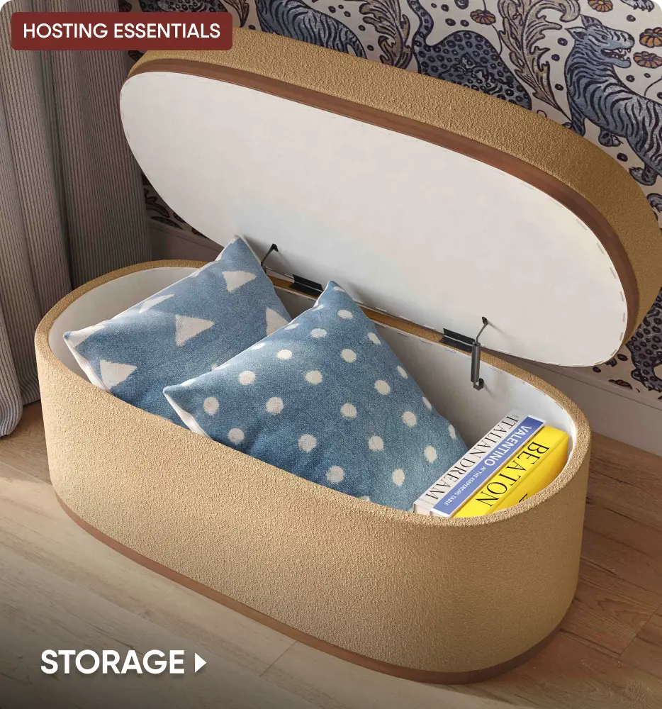 HOSTING ESSENTIALS STORAGE: An oval, tan textured storage ottoman with its lid open, revealing two pillows and books inside.