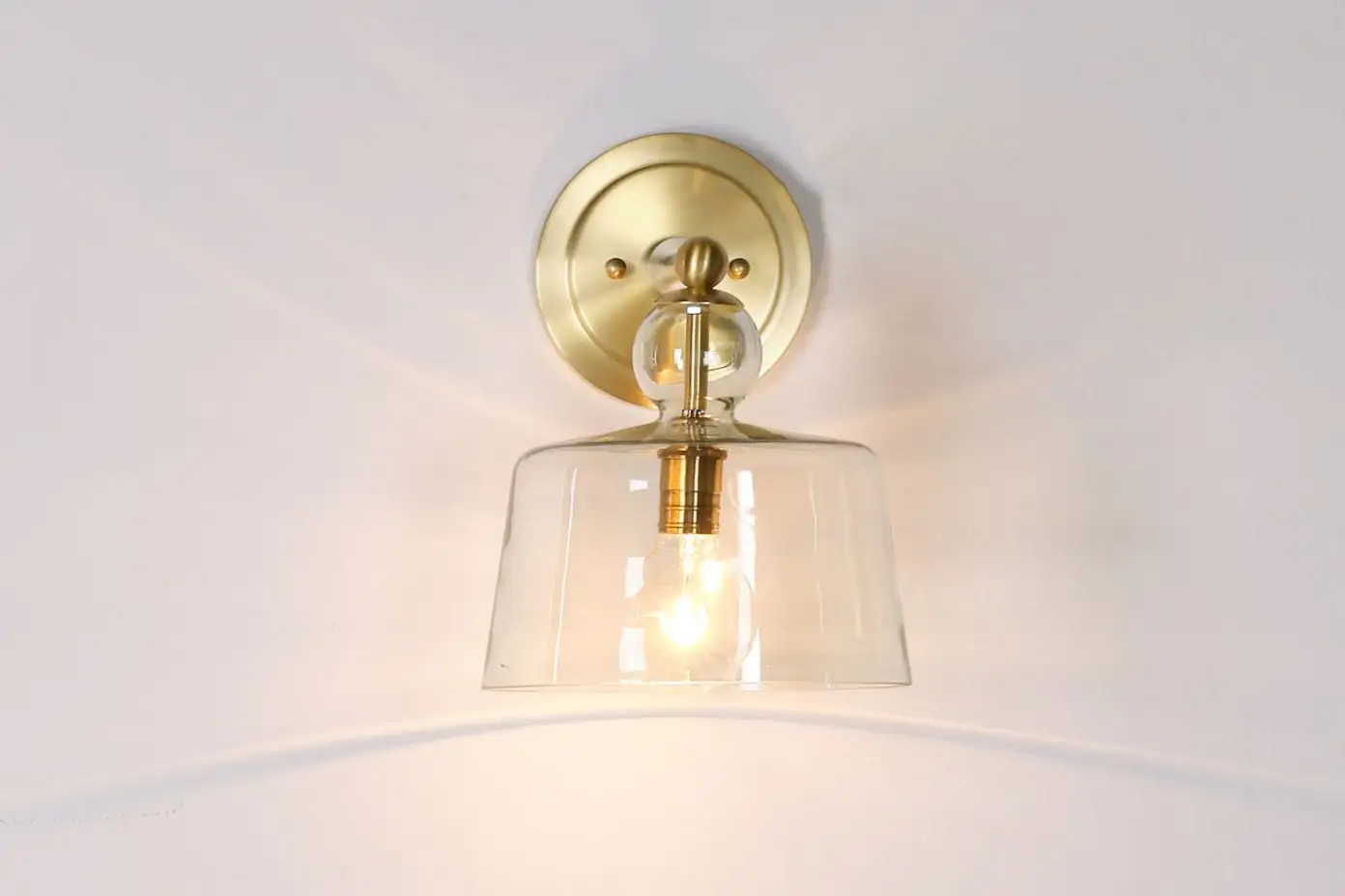 A brass wall sconce with a clear glass shade, illuminating a white wall.
