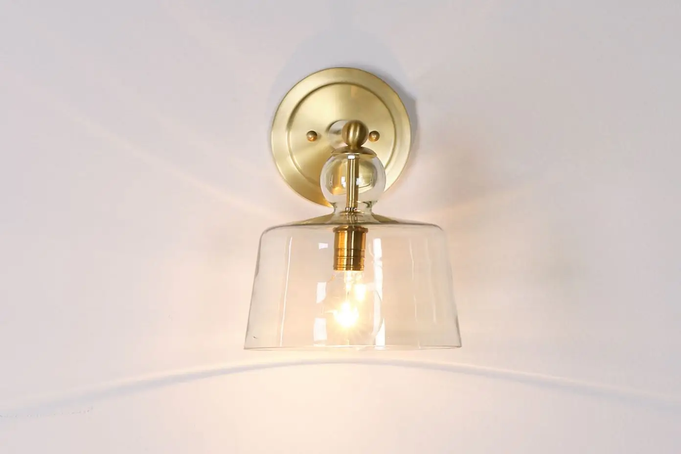 A brass wall sconce with a clear glass shade, illuminating a white wall.