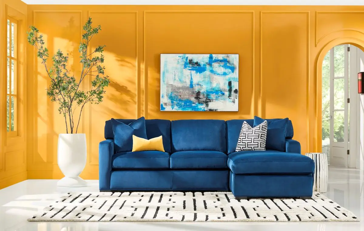 A blue velvet sectional sofa with a chaise, accented by yellow and patterned pillows, against a vibrant yellow wall.