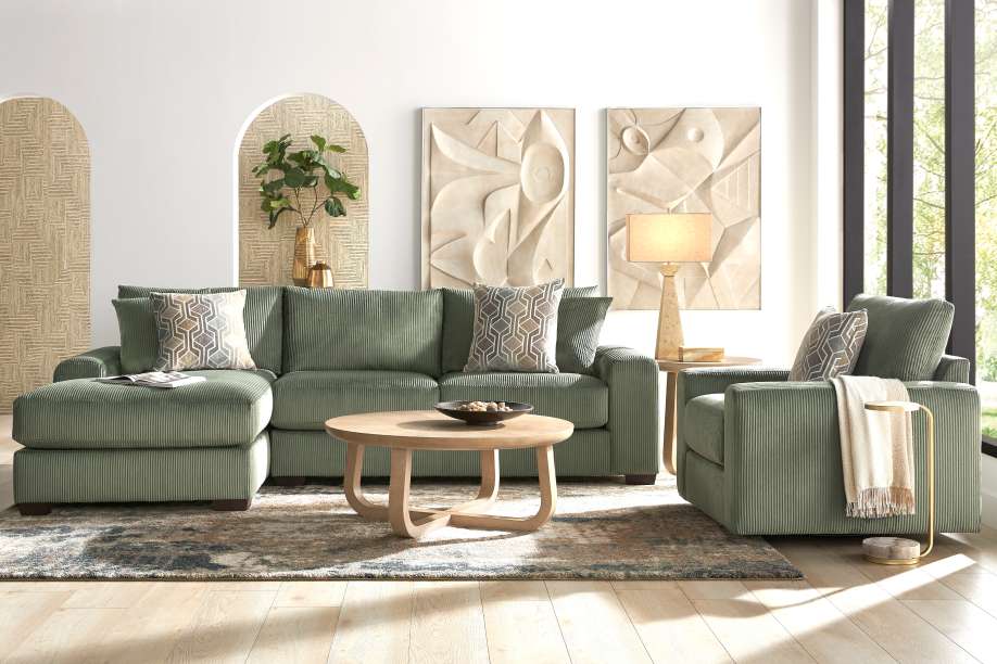 Green corduroy sectional sofa with matching armchair, round wooden coffee table, and abstract wall art.
