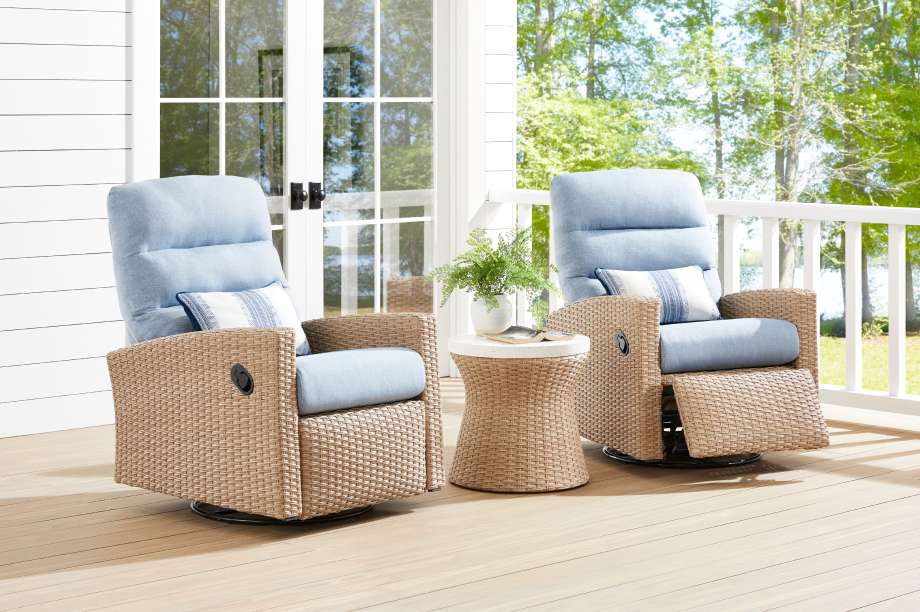 Two wicker outdoor recliner chairs with blue cushions on a wooden deck. A small side table with a plant is between them.
