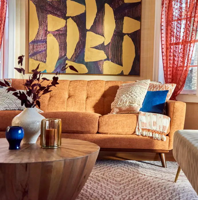 An orange sofa with decorative pillows and a throw blanket, positioned in front of a large abstract painting.