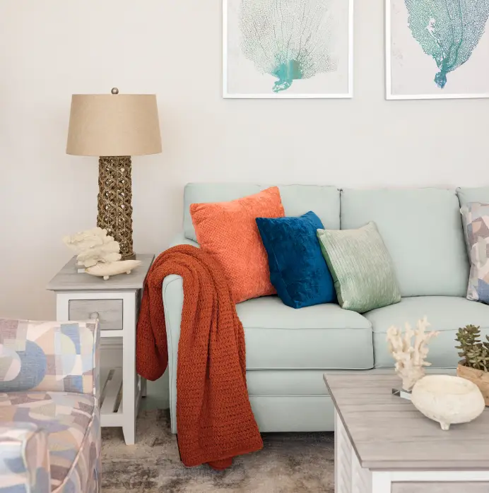 A light blue sofa with orange, blue, and green pillows, a rust-colored throw blanket, and a side table with a lamp.