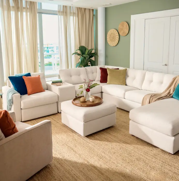 A cream-colored sectional sofa with colorful throw pillows and a matching ottoman in a bright, modern living room.