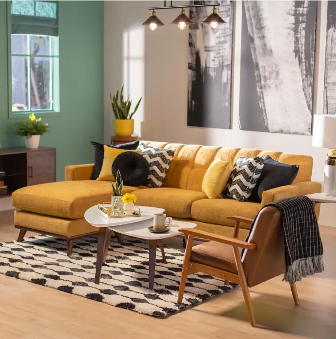 A mustard yellow sectional sofa with patterned pillows, a white coffee table, and a brown armchair on a patterned rug.