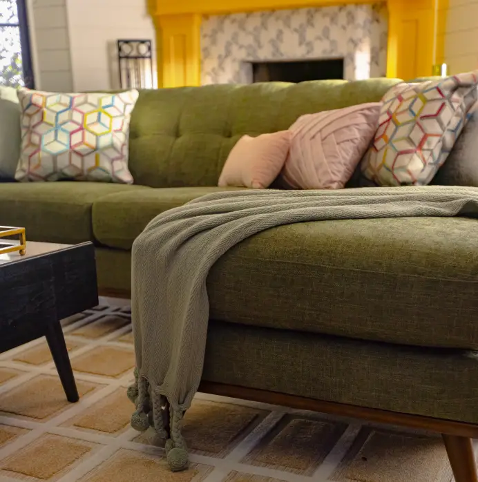 A green mid-century modern sofa with patterned and solid throw pillows and a draped throw blanket on a patterned rug.