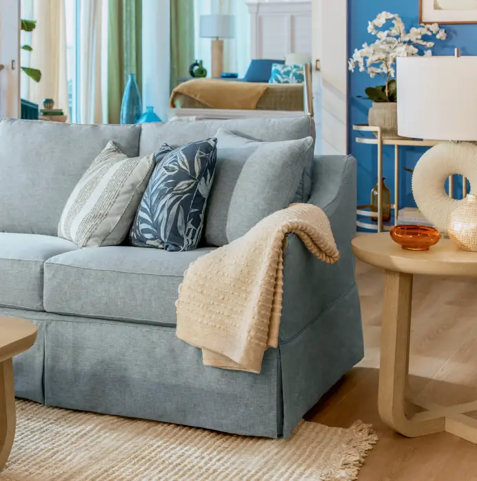 A light blue sofa with patterned throw pillows and a beige knit throw blanket, next to a round side table with a lamp.