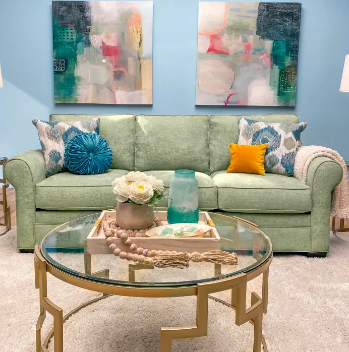 A light green sofa with patterned and solid accent pillows sits in front of two abstract paintings and a gold coffee table.
