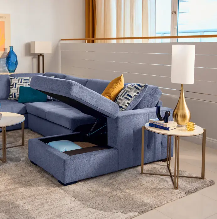 A blue sectional sofa with a lift-up storage compartment, featuring decorative pillows and a side table with a lamp.