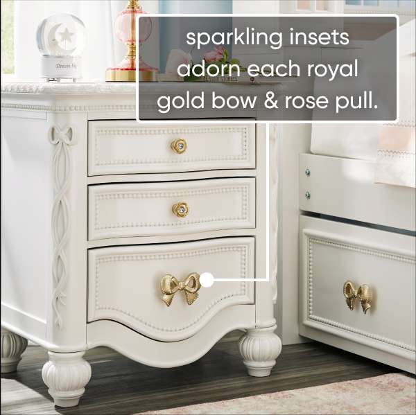 sparkling insets adorn each royal gold bow & rose pull. A white nightstand with gold hardware.
