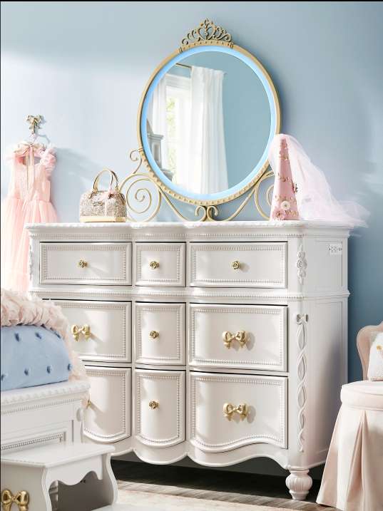 White dresser with gold bow drawer pulls, a round mirror, and a pink dress.
