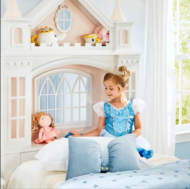 A young girl in a princess dress sits on a bed with a castle-shaped headboard.
