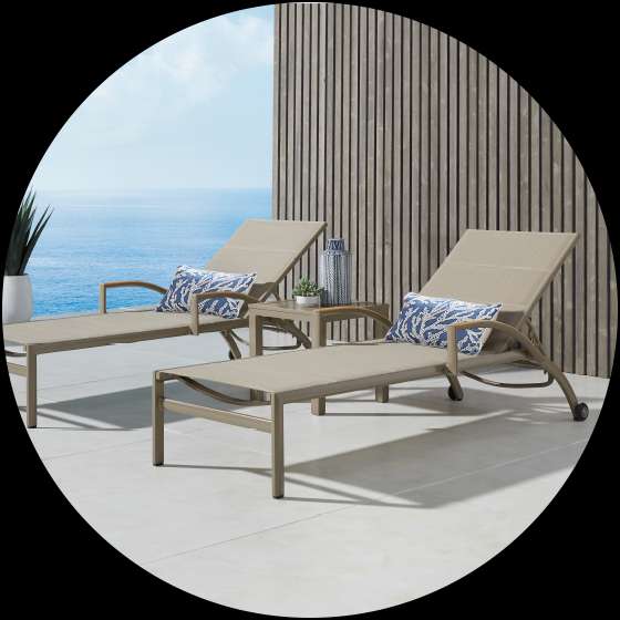 Two beige chaise lounges with blue pillows on a patio overlooking the ocean.

