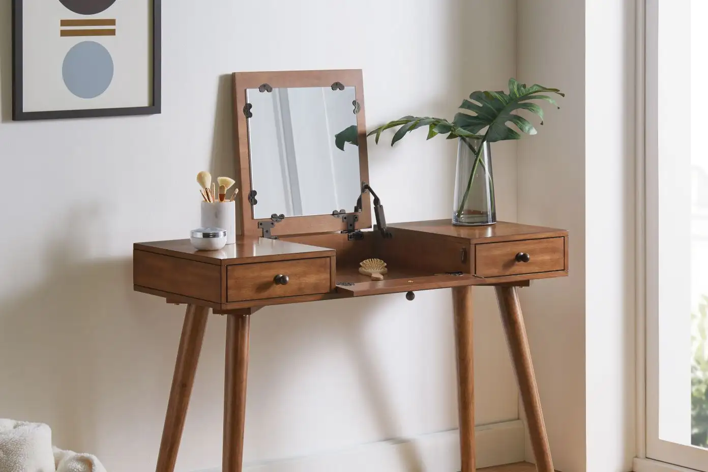 Wooden vanity table with a mirror, drawers, and a vase of leaves.
