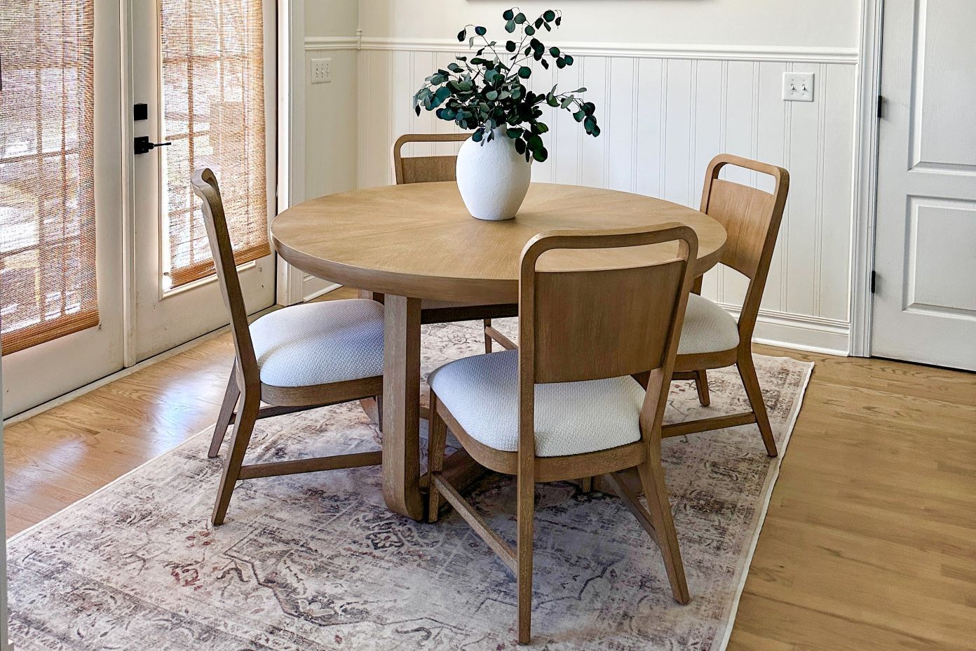 A round wooden dining table with four chairs on a patterned rug. A vase with greenery sits on the table.
